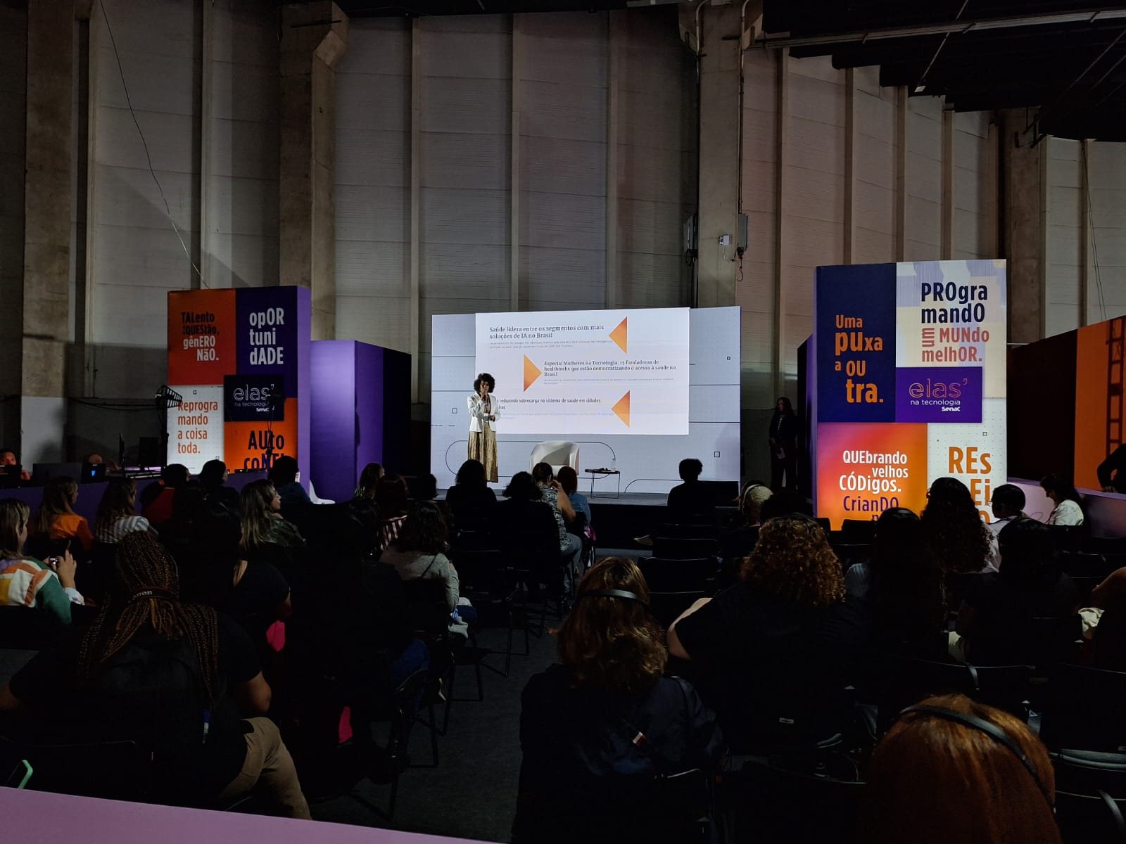 Mariza Faria no palco de um auditório, com projeção de conteúdo, diante do público sentado.